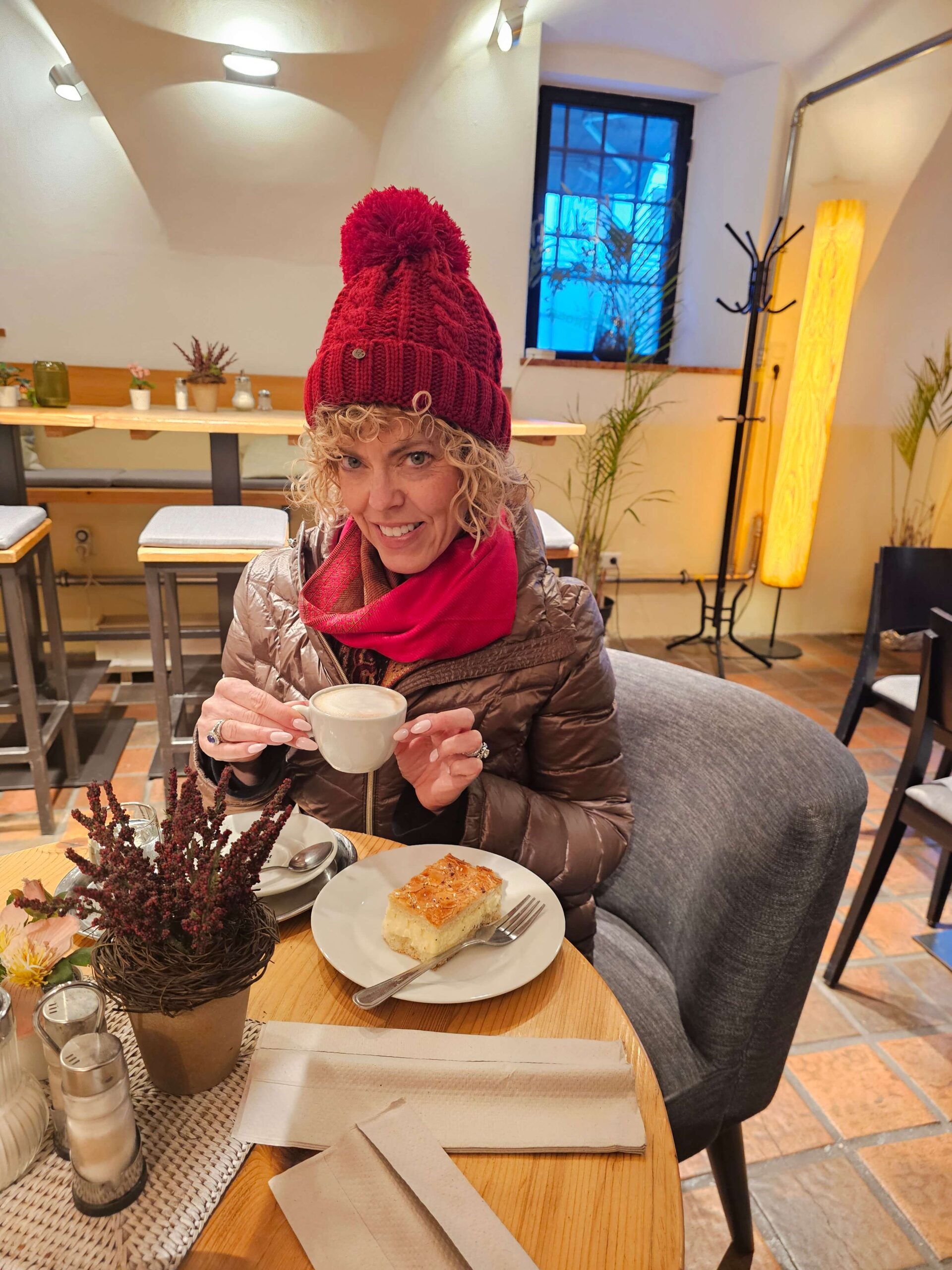 lady sitting in coffee shop sipping coffee in red scarf coat and pom pom hat: for blog post "are river cruises worth it"