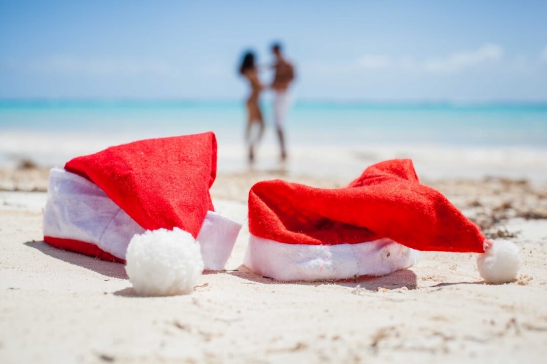 2 santa hats laying on the beach with a couple in the background. for the blog, best travel gifts for Christmas