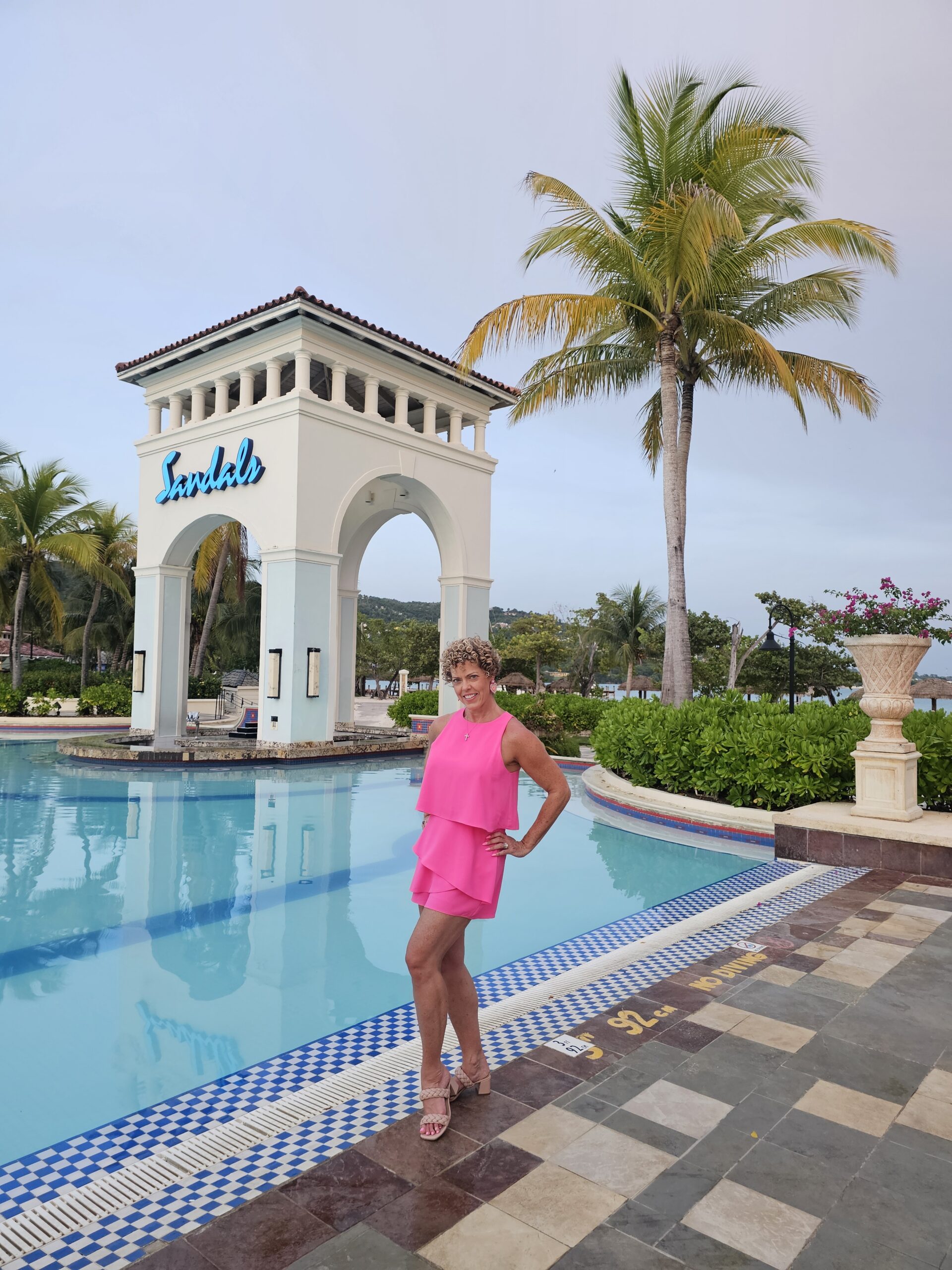 woman in pink shorts jumpsuit standing in front of a pool and large archway saying Sandals; from blog post Best Sandals Resorts