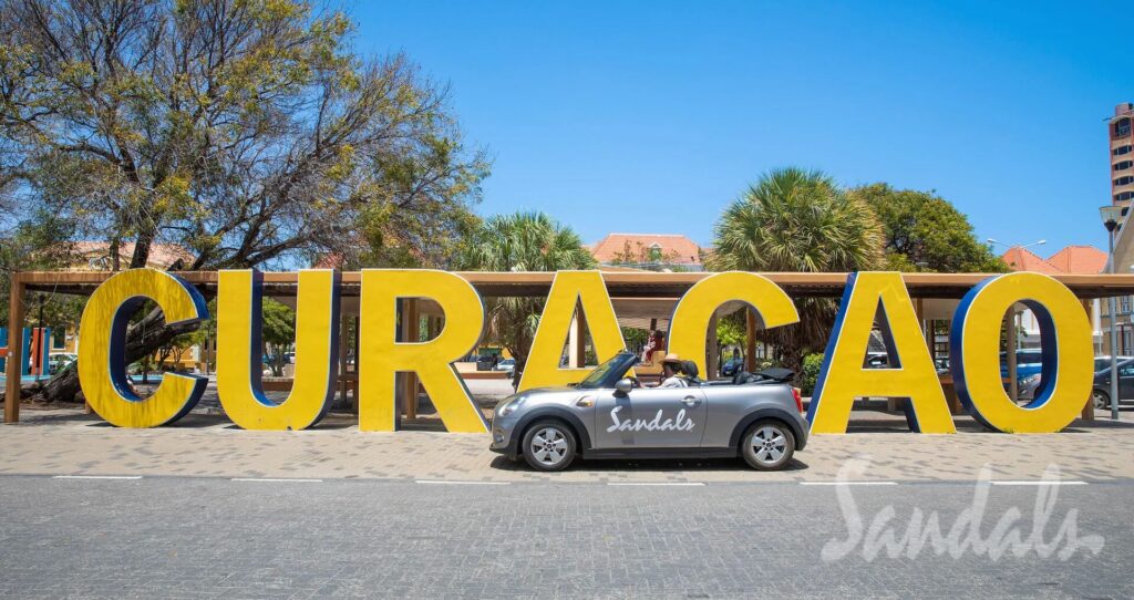 big sign that says Curacao and a silver Mini Cooper in front of it